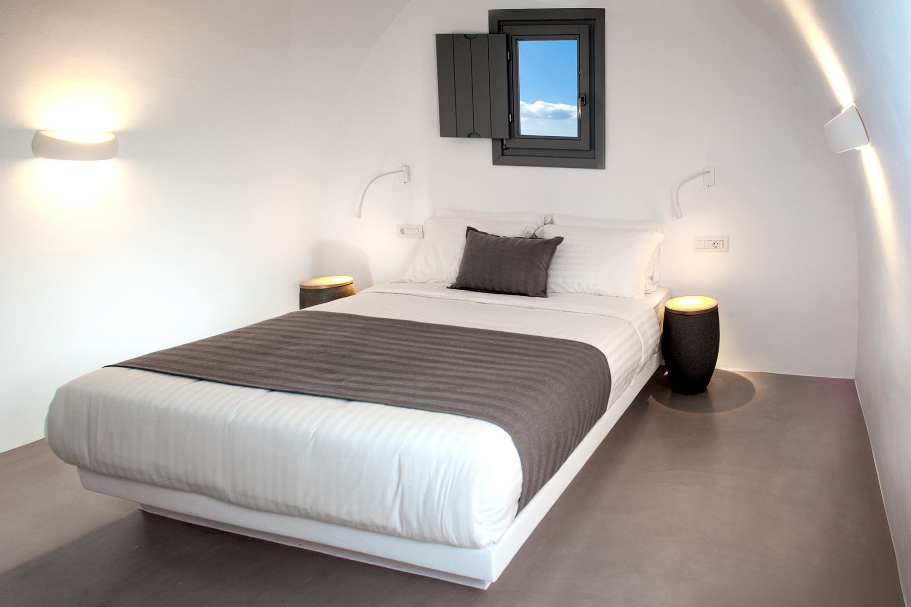 A modern bedroom with a large bed, two bedside tables, a window with a view, and wall-mounted lights.