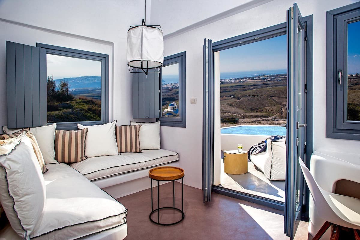 Una sala de estar moderna con un sofá, una mesita redonda y ventanas amplias con vistas a un paisaje costero.