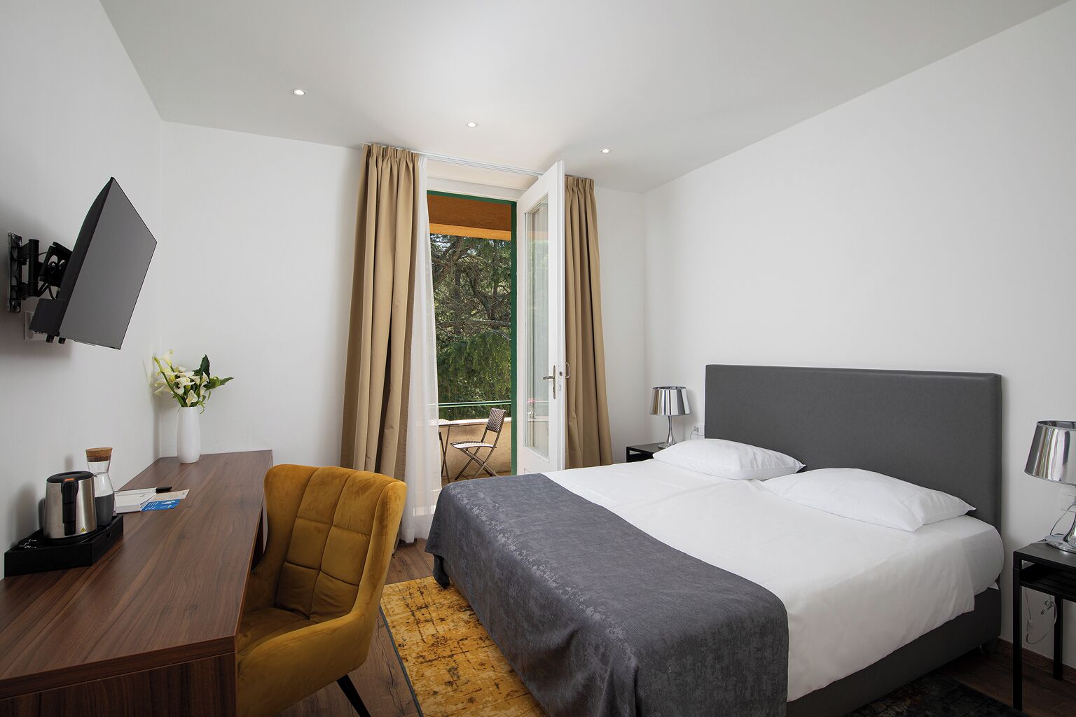 A hotel room with a bed, a desk with a chair, a TV mounted on the wall, and a view of greenery outside.