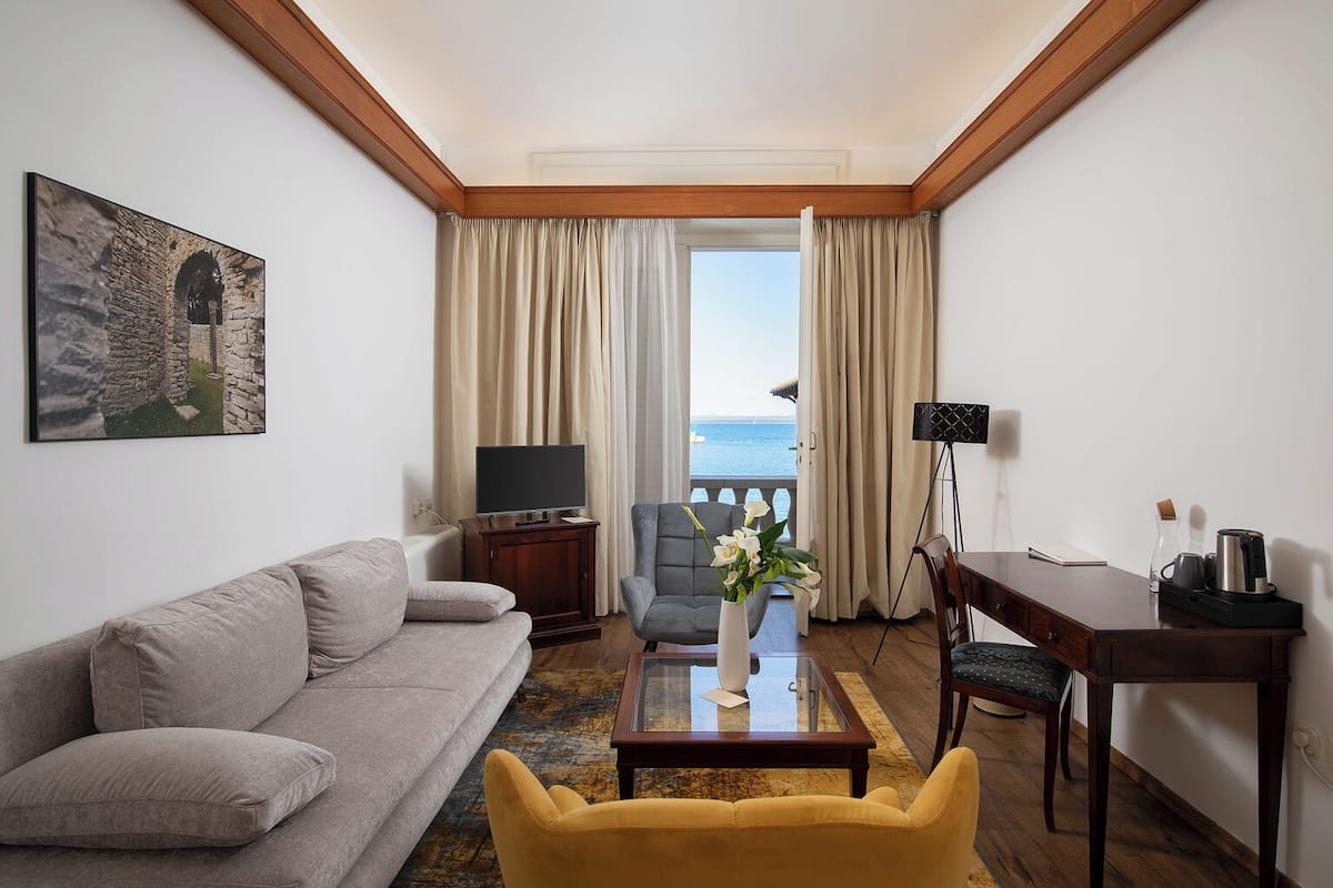 A hotel room with a sofa, armchair, coffee table, TV, and a view of the sea.