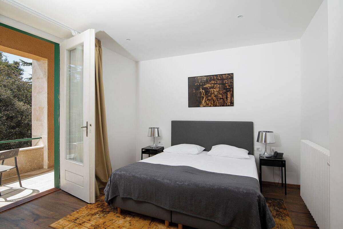 A bedroom with a bed, two bedside tables, a radiator, a door leading to a balcony, and a painting on the wall.