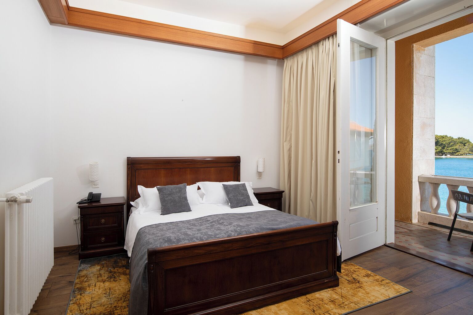A bedroom with a wooden bed, a bedside table, a radiator, and a view of the sea through a balcony.