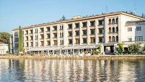A waterfront hotel with multiple balconies, outdoor seating, and a reflection in the water. - Brijuni Hotel Neptun (Pula)