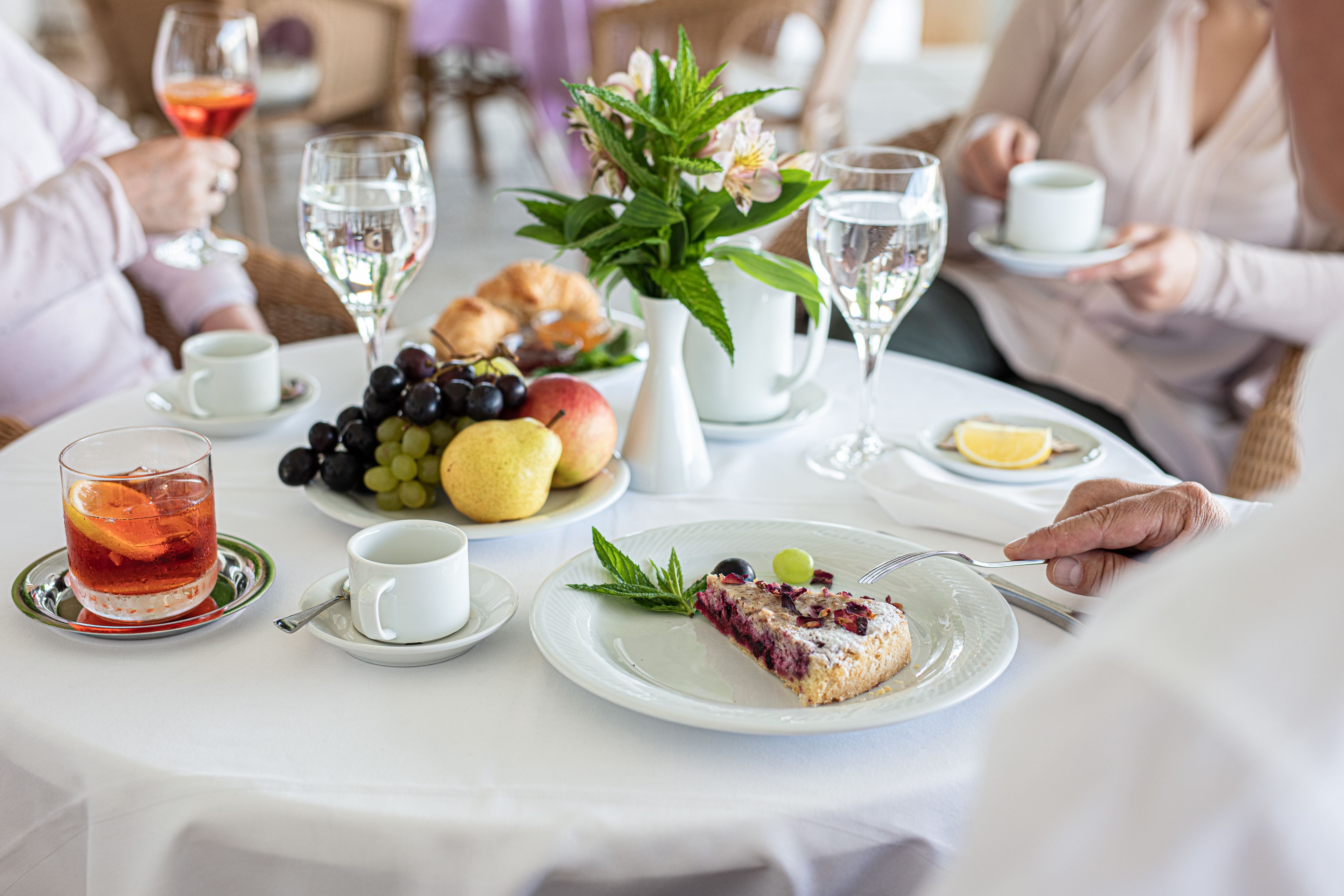 A dining table with a variety of fruits, a slice of cake, and glasses of water and tea.