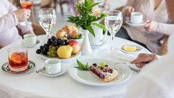 A dining table with a variety of fruits, a slice of cake, and glasses of water and tea.