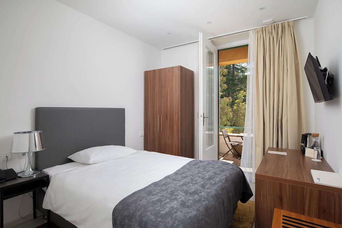 A hotel room with a bed, a desk, a TV, and a balcony with curtains.
