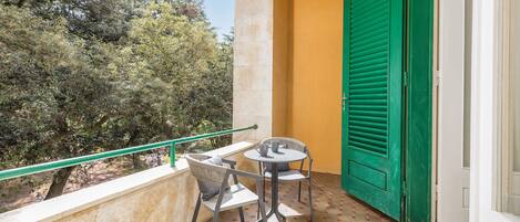 Een balkon met een tafel en stoelen, een groene deur en uitzicht op bomen.