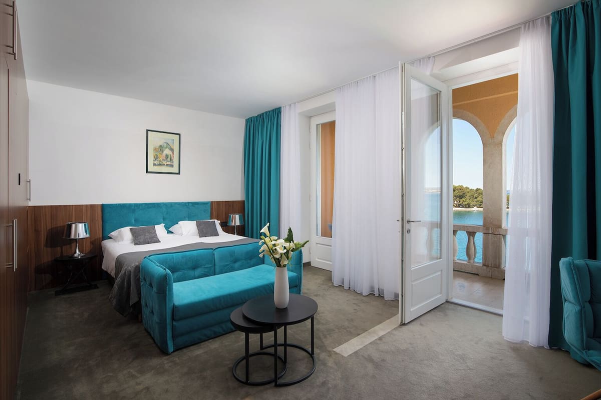 A hotel room with a bed, a small table with a vase, a nightstand, and a view of the sea through a balcony.