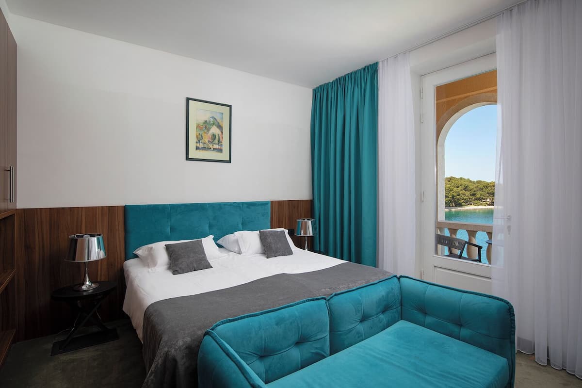 A hotel room with a bed, a sofa, a window with a view, a painting on the wall, and a nightstand with a lamp.