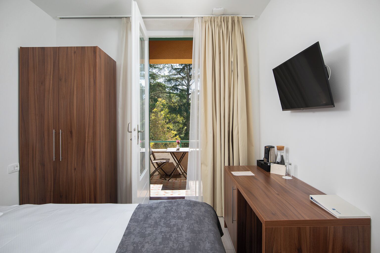 A hotel room with a bed, a desk, a TV, and a balcony with curtains.