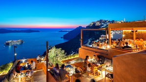 A rooftop dining area with tables and chairs, overlooking a coastal cityscape and the sea. - Athina Aperto Suites (Santorini)