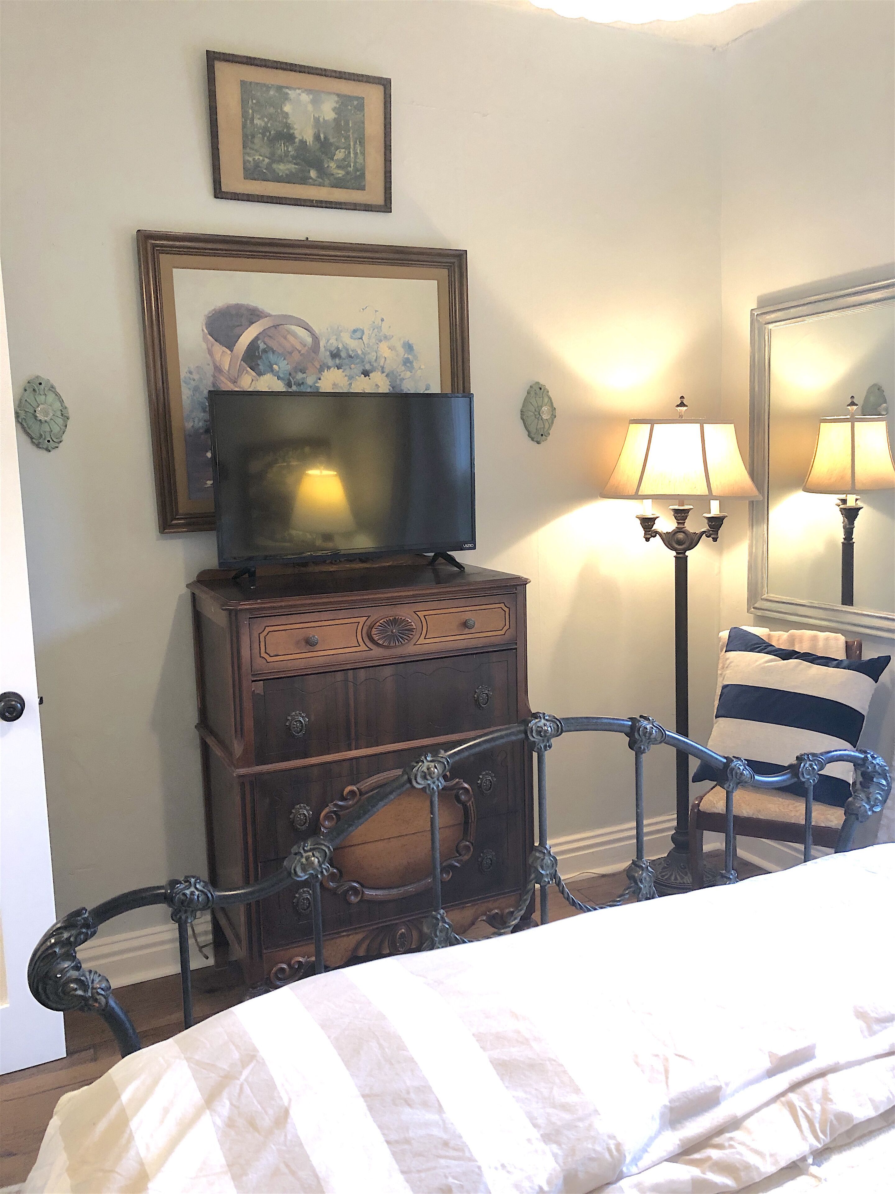 Ein Schlafzimmer mit einem Bett, einer Kommode mit Fernseher darauf, einer Lampe und einem Sessel mit gestreiftem Kissen.