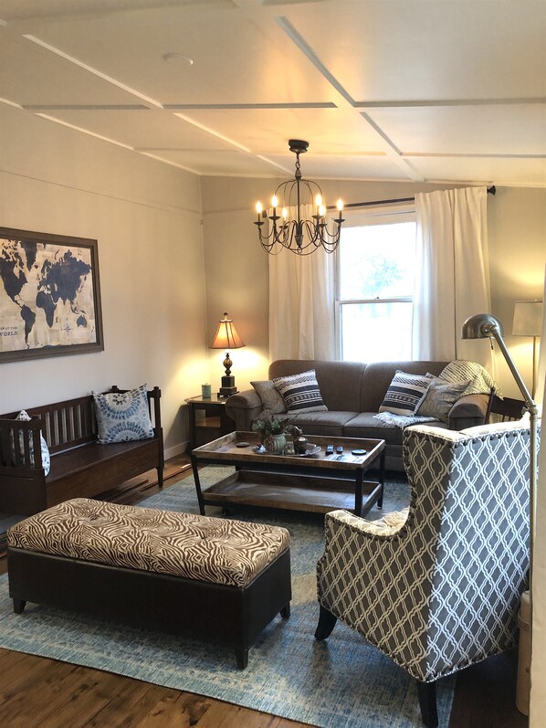 A living room with a sofa, coffee table, patterned ottoman, and armchair. A world map is on the wall, and a chandelier hangs from the ceiling.