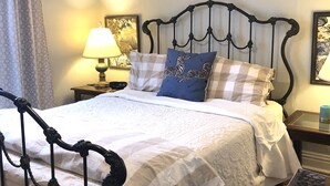 Een net opgemaakt bed met een geruit sprei, een blauw kussen met zebra’s en een wit kussen. Boven het bed hangt een krans aan de muur, een lamp op een nachtkastje en een lijst met een afbeelding aan de muur.