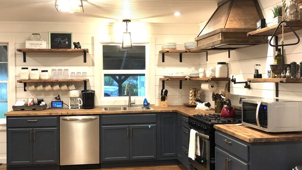 Een moderne keuken met donkere kastjes, een RVS vaatwasser, een gasfornuis en een magnetron. Daarboven staan houten planken met diverse keukenspullen.