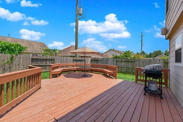 Une terrasse en bois avec un barbecue et une table, entourée d’une clôture en bois et avec des maisons en arrière-plan.