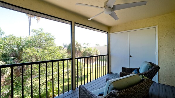 A balcony with wicker furniture, a ceiling fan, and a view of a garden with various plants.