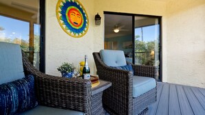 A covered outdoor seating area with wicker chairs, a small table with a bottle and glasses, and a decorative sun mural on the wall. - Coastal Condo, Heated Pool Steps from the Beach (Holmes Beach)
