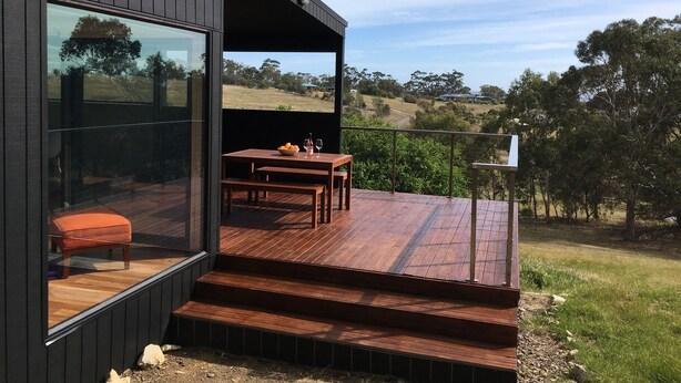 A modern outdoor deck with a wooden table and bench, a glass door leading to a room, and a scenic view of a rural landscape. - Architectural Design Rural Retreat (Forcett)