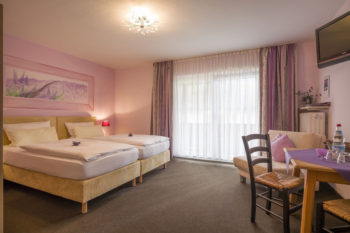 A hotel room with two beds, a dining table with chairs, a radiator, and a painting on the wall.