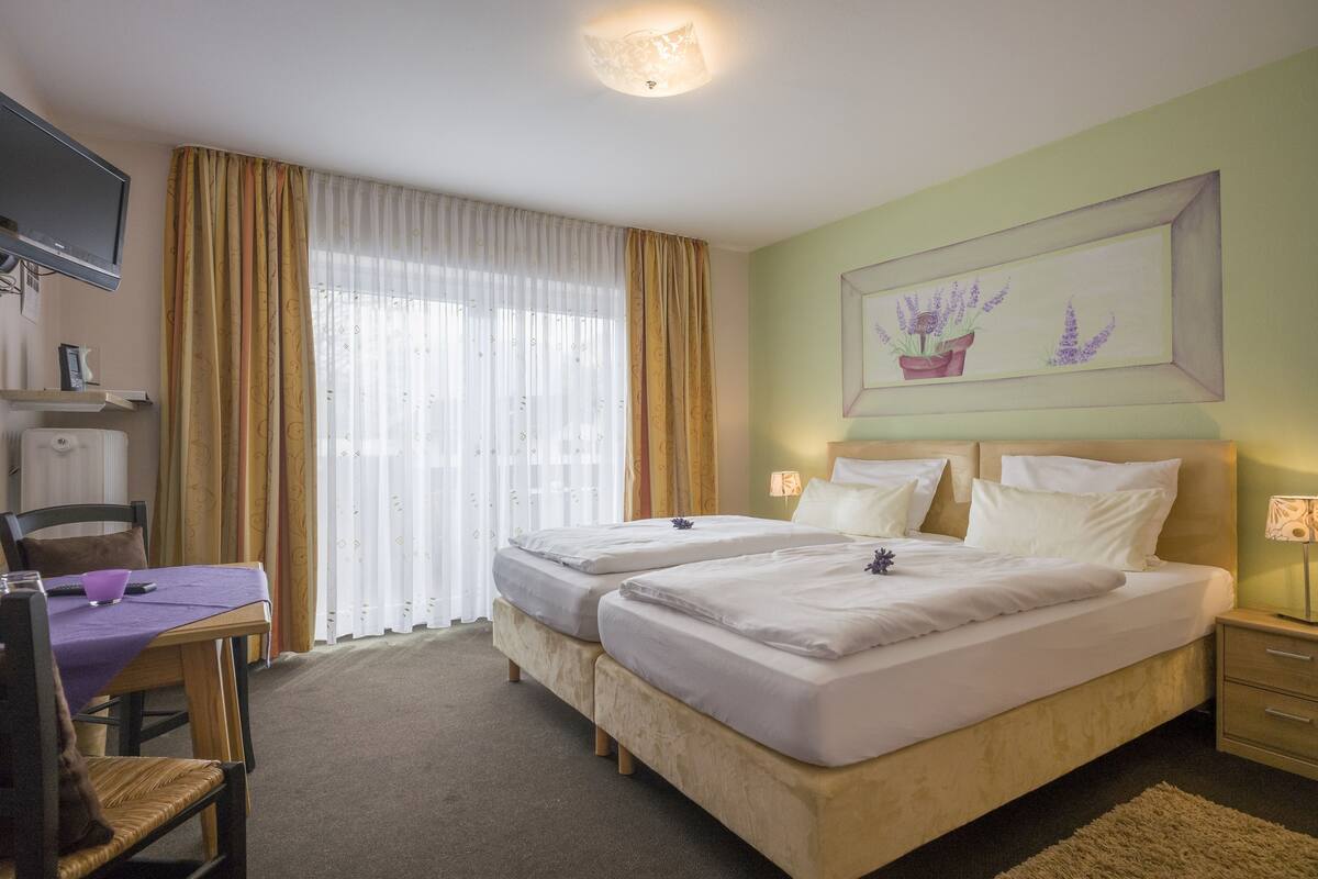 A hotel room with two beds, a desk with a purple tablecloth, a chair, a television, and a window with curtains.