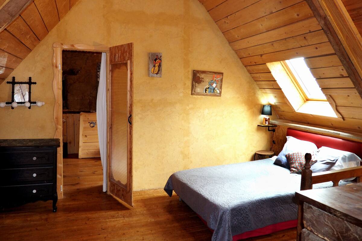 Un acogedor dormitorio en el ático con techo inclinado, paredes de madera, una cama con cabecera roja, una mesita de noche con una lámpara y una ventana con vistas al cielo.