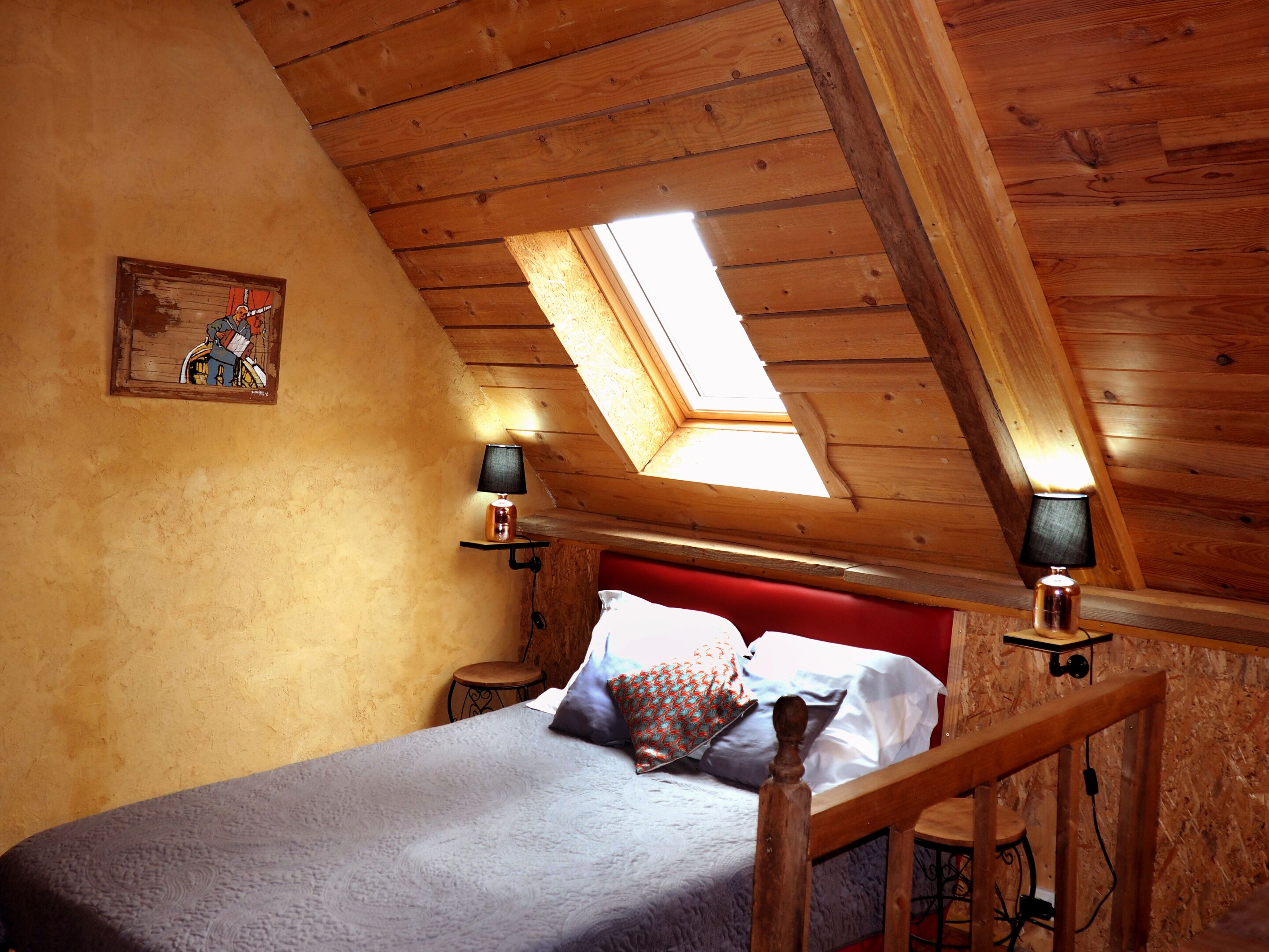A bedroom with a sloped ceiling, a skylight, a bed with a red headboard, two bedside lamps, and a small round table.
