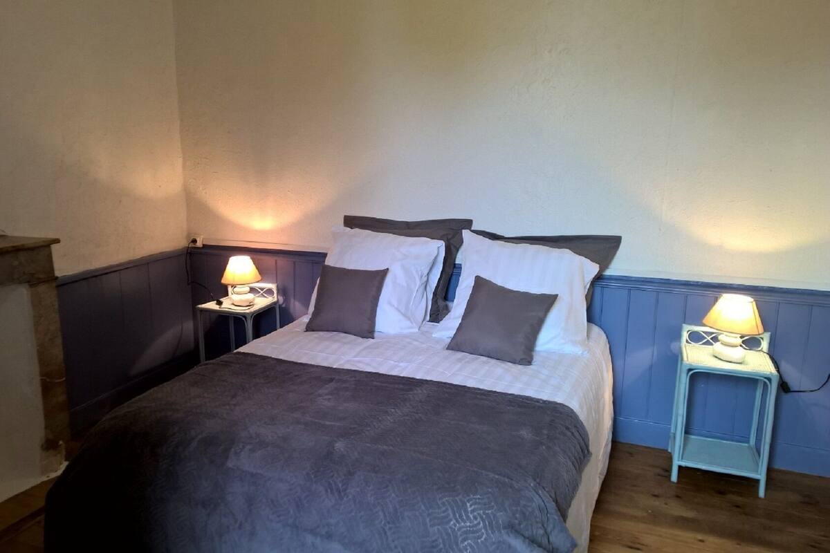 Un dormitorio con cama, dos mesitas de noche con lámparas, cabecera de madera y un taburete pequeño.