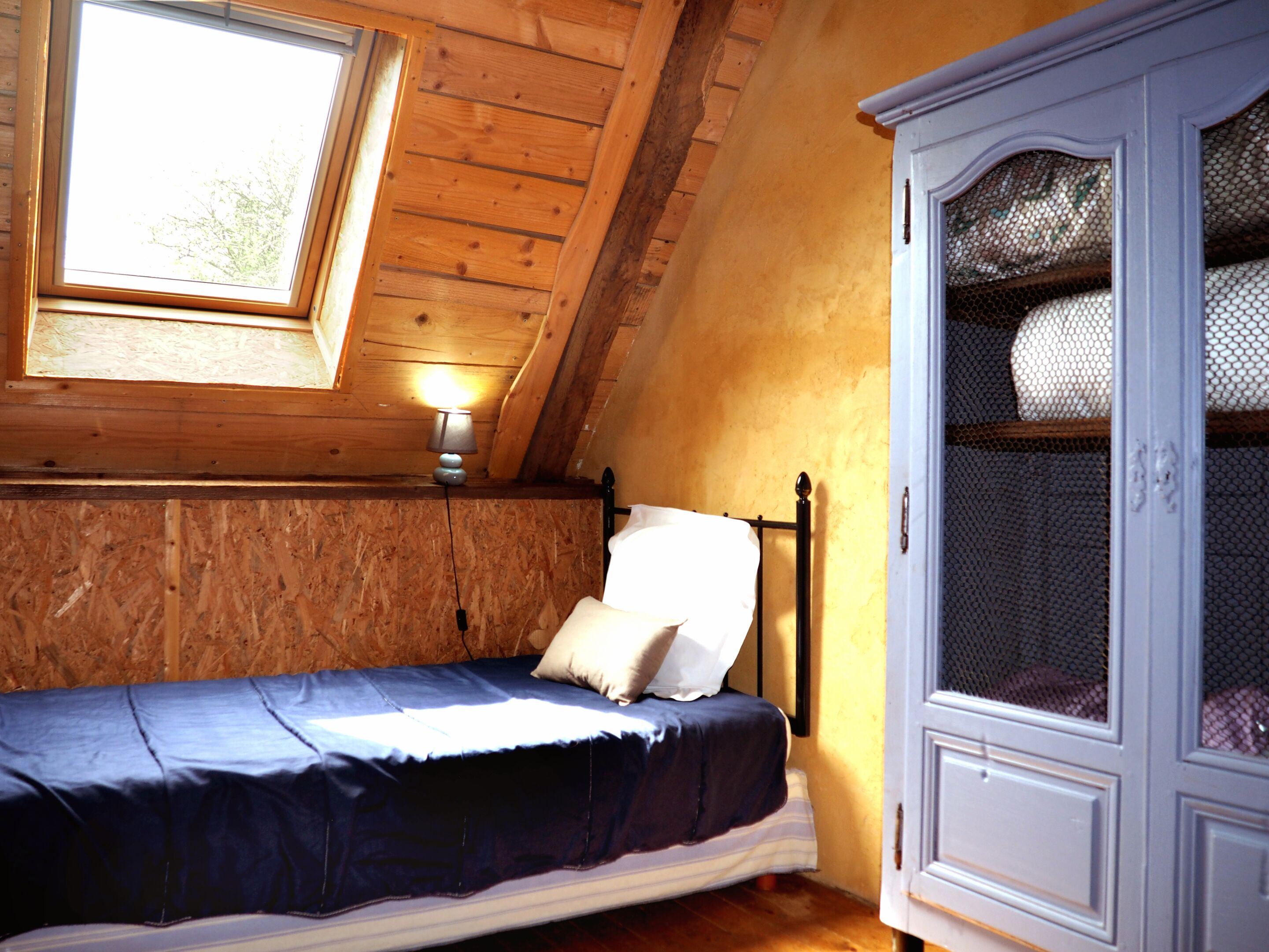 Una cama individual con cabecera, una ventana y un armario en una habitación con paredes de madera y techo inclinado.