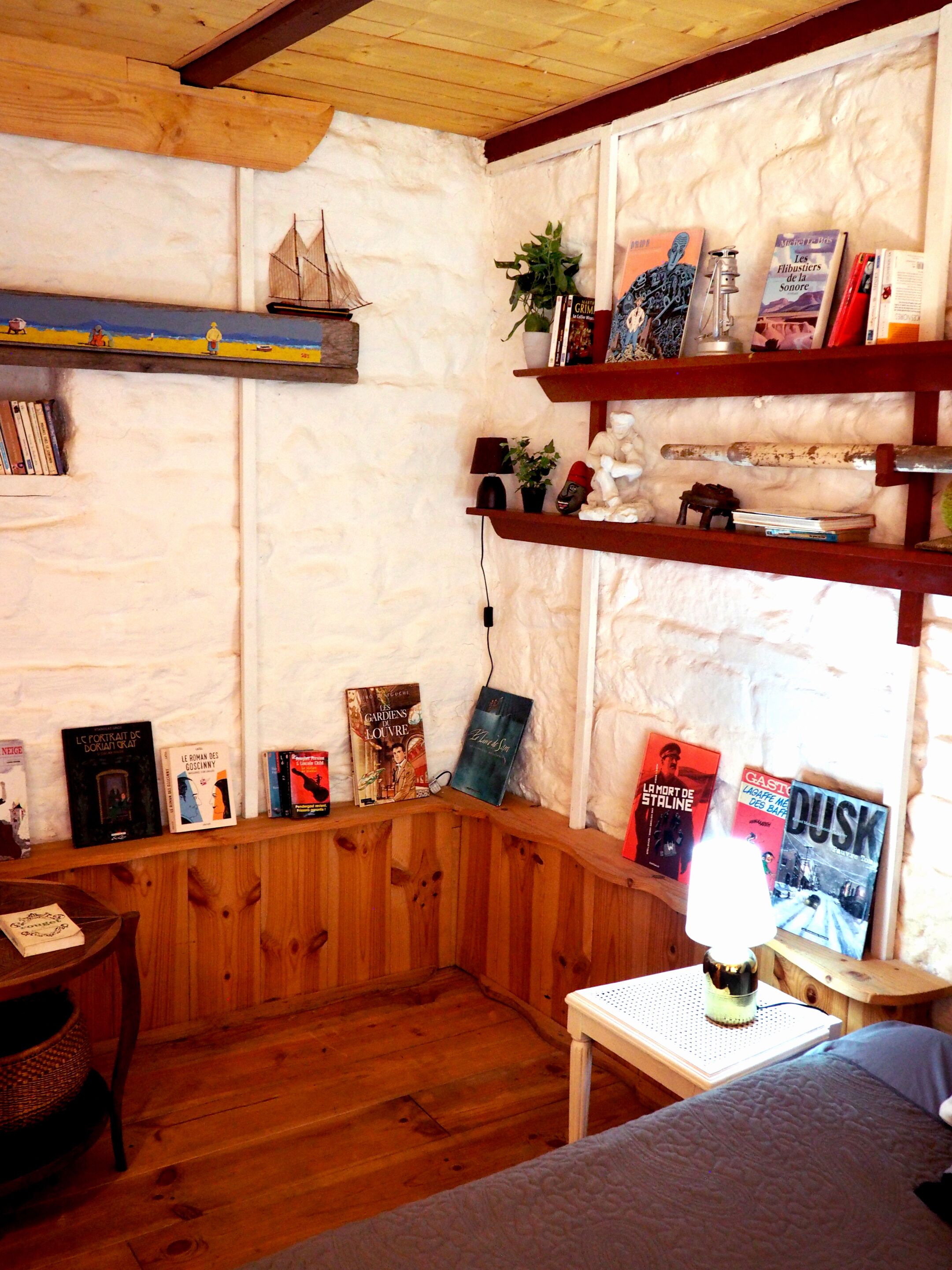 Una habitación acogedora con paredes de madera y techo inclinado. Se ven estanterías con libros y adornos, una mesita con una lámpara y un banco.