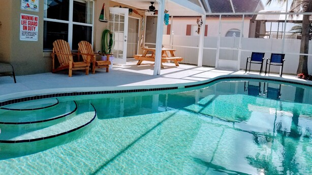 A pool area with clear water, wooden chairs, and a covered patio. - The Cozy House (Kissimmee)