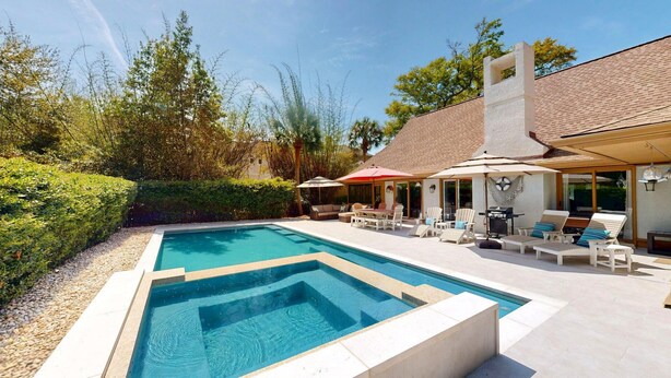 Ein Swimmingpool mit abgesenktem Bereich, umgeben von einer Terrasse mit Liegestühlen und Sonnenschirmen, sowie ein Haus mit Ziegeldach und großen Fenstern.