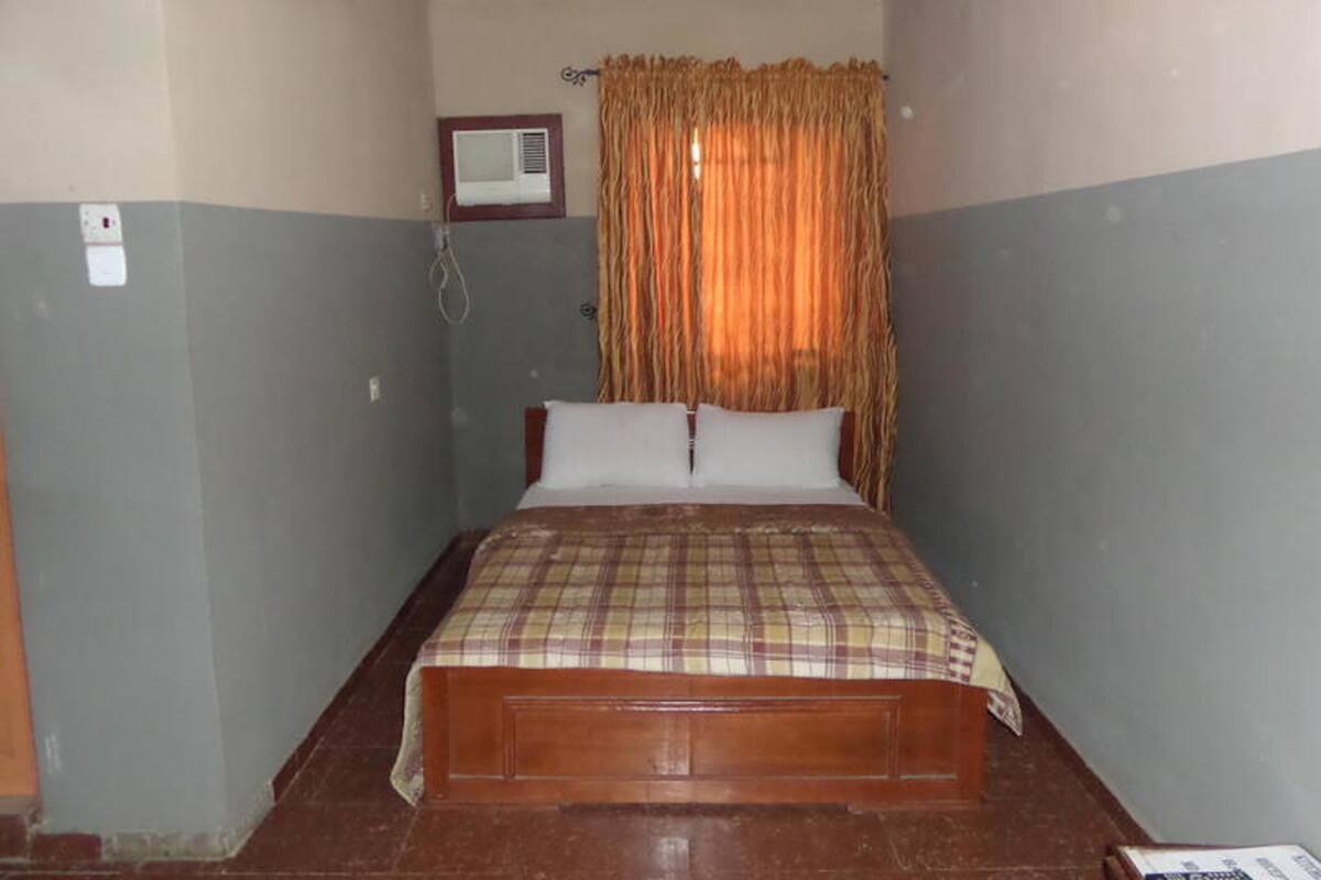 A small, single-bed room with a wooden headboard, a ceiling fan, an air conditioning unit, and a curtain.