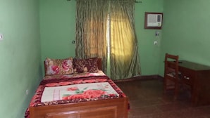 A bedroom with a wooden bed, green walls, a window with curtains, a desk, and an air conditioning unit.