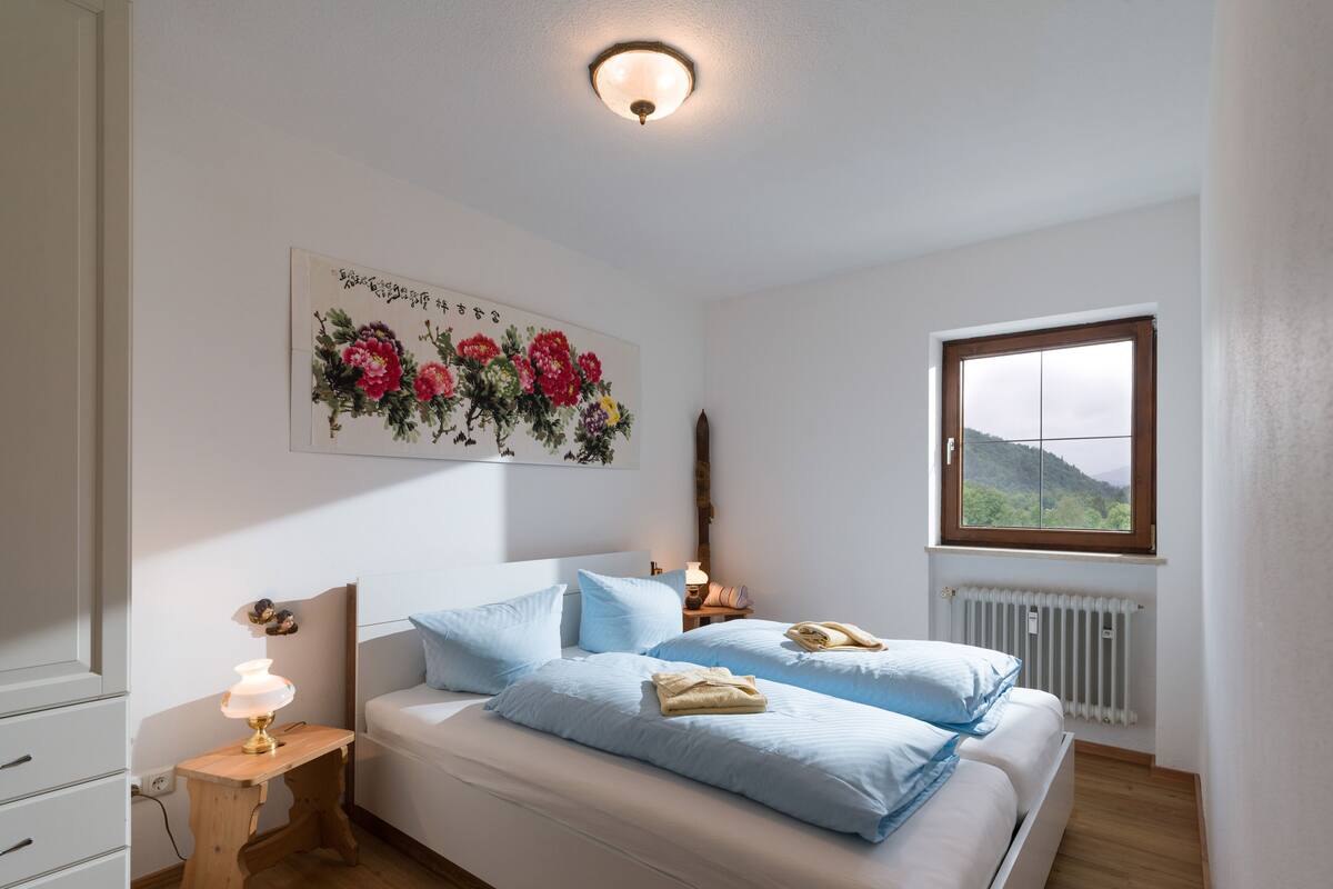 Een slaapkamer met een bed, nachtkastje, lamp en een raam met uitzicht op groen.
