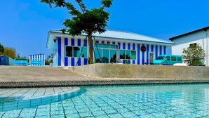 Una piscina con borde de azulejos, un edificio con franjas verticales azules y blancas, y una furgoneta turquesa estacionada.