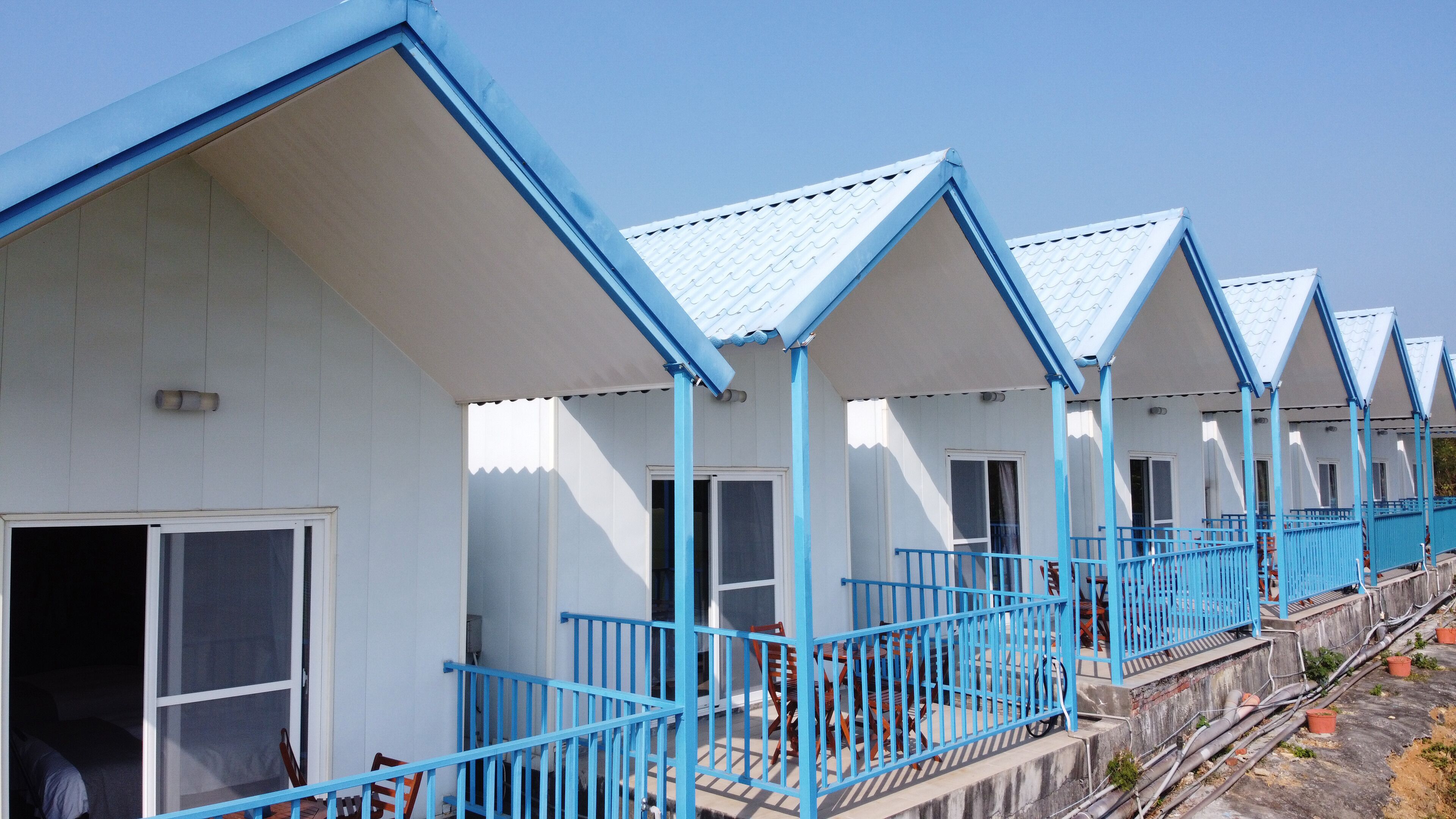 A row of beach huts with blue railings and white walls, featuring balconies and a clear sky.