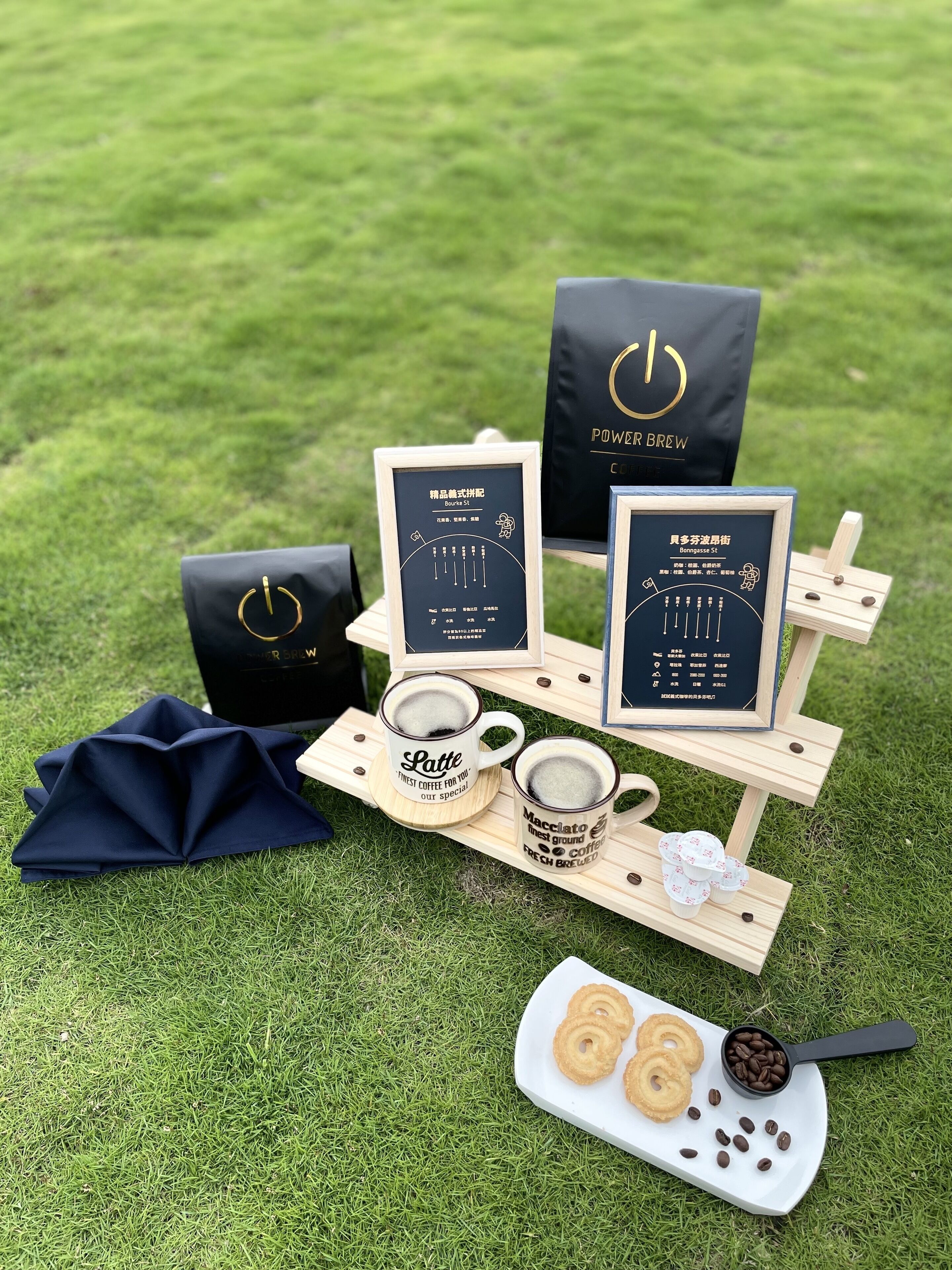 A coffee tasting setup with two mugs labeled 'Latte' and 'Macchiato', a tray with coffee beans and pastries, and branded bags on a grassy surface.