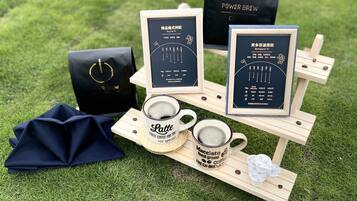 A coffee tasting setup with two mugs labeled 'Latte' and 'Macchiato', a tray with coffee beans and pastries, and branded bags on a grassy surface.
