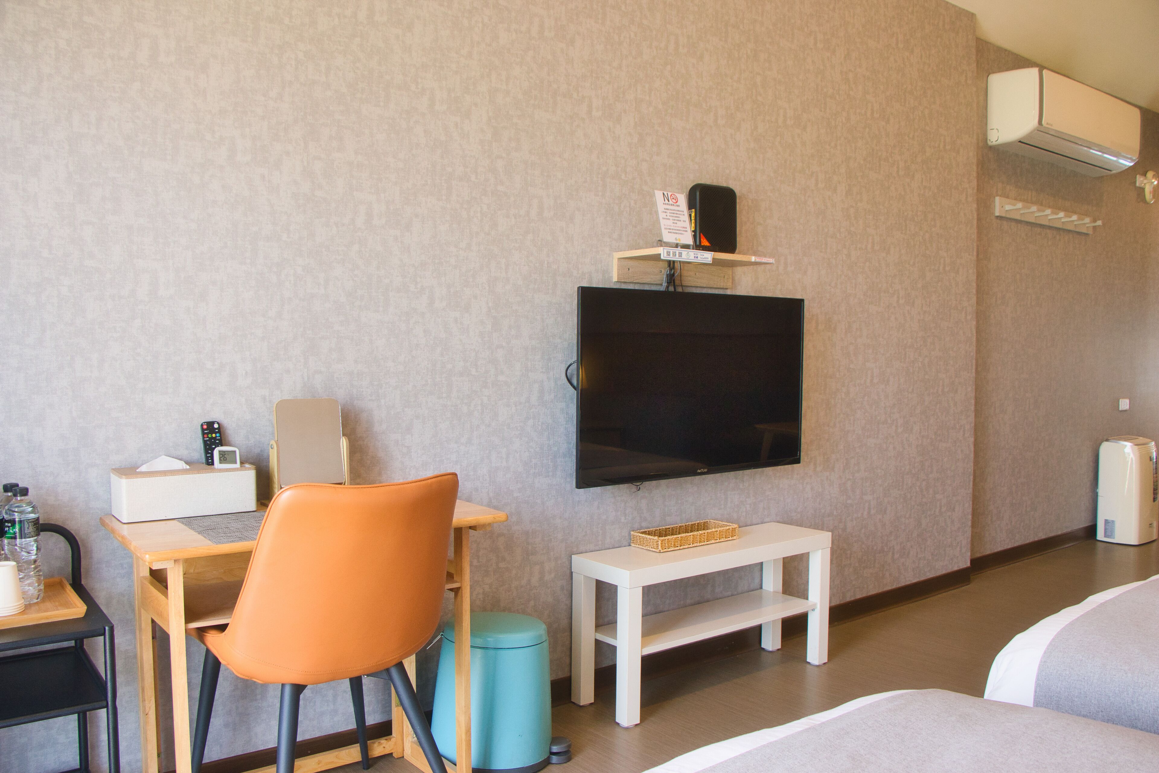 A hotel room with a bed, a desk with a chair, a television mounted on the wall, a small table, and a trash bin.