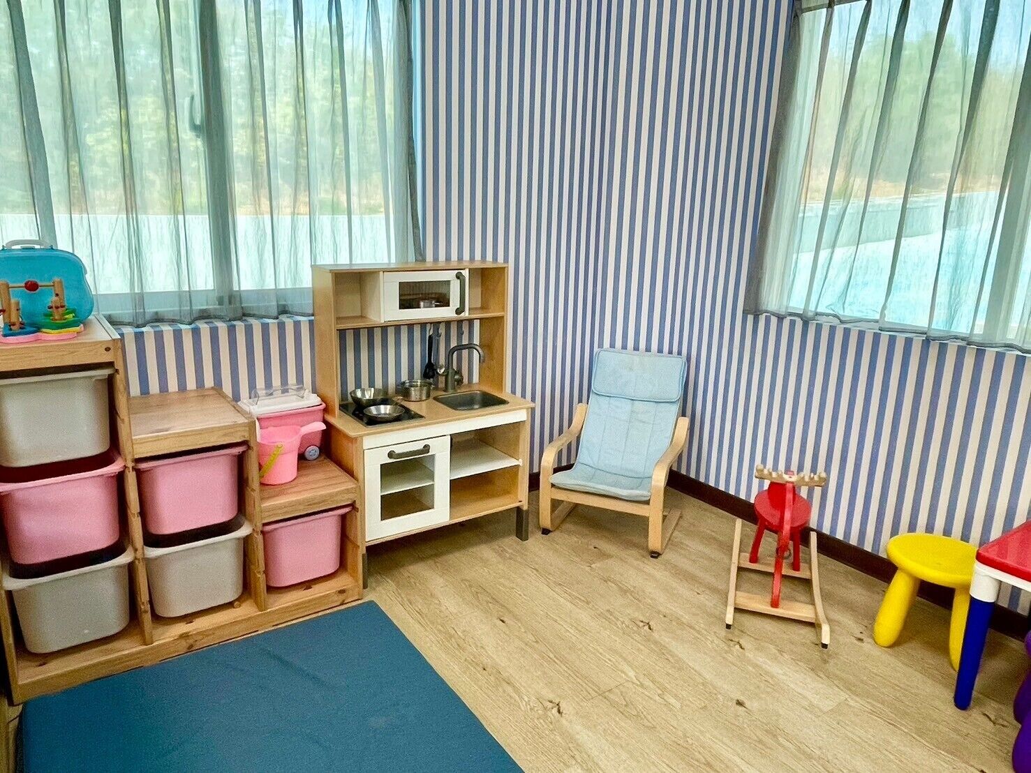 A children's play area with a play kitchen, storage bins, a blue mat, and a window with curtains.