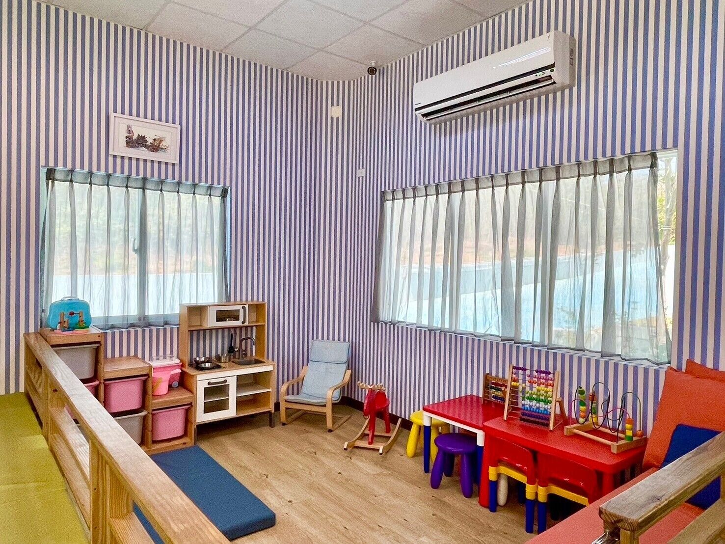 A children's playroom with striped walls, a wooden bench, a rocking chair, and a red table with toys.
