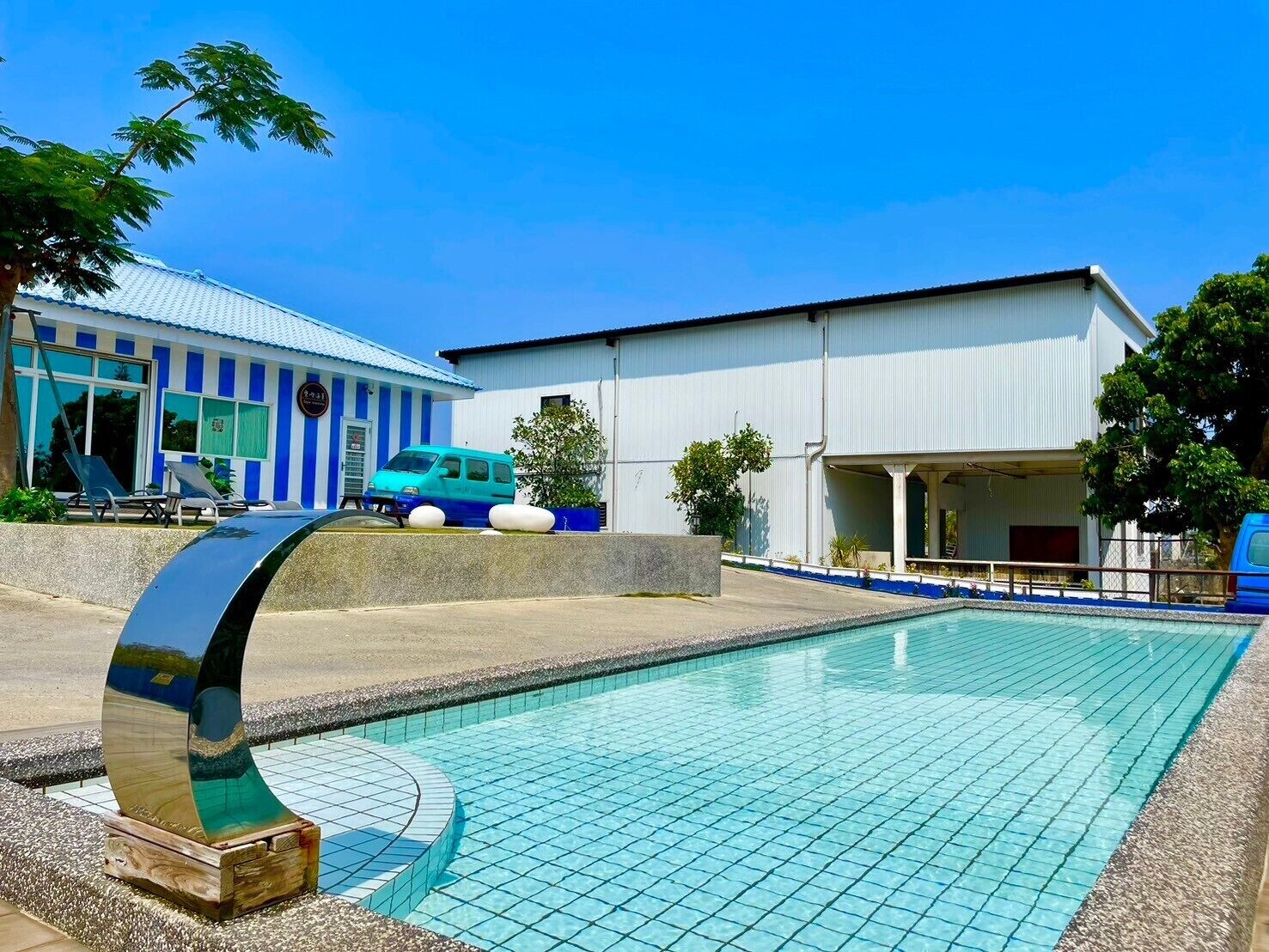 A swimming pool with a tiled deck, a blue van, and a building with blue and white stripes.