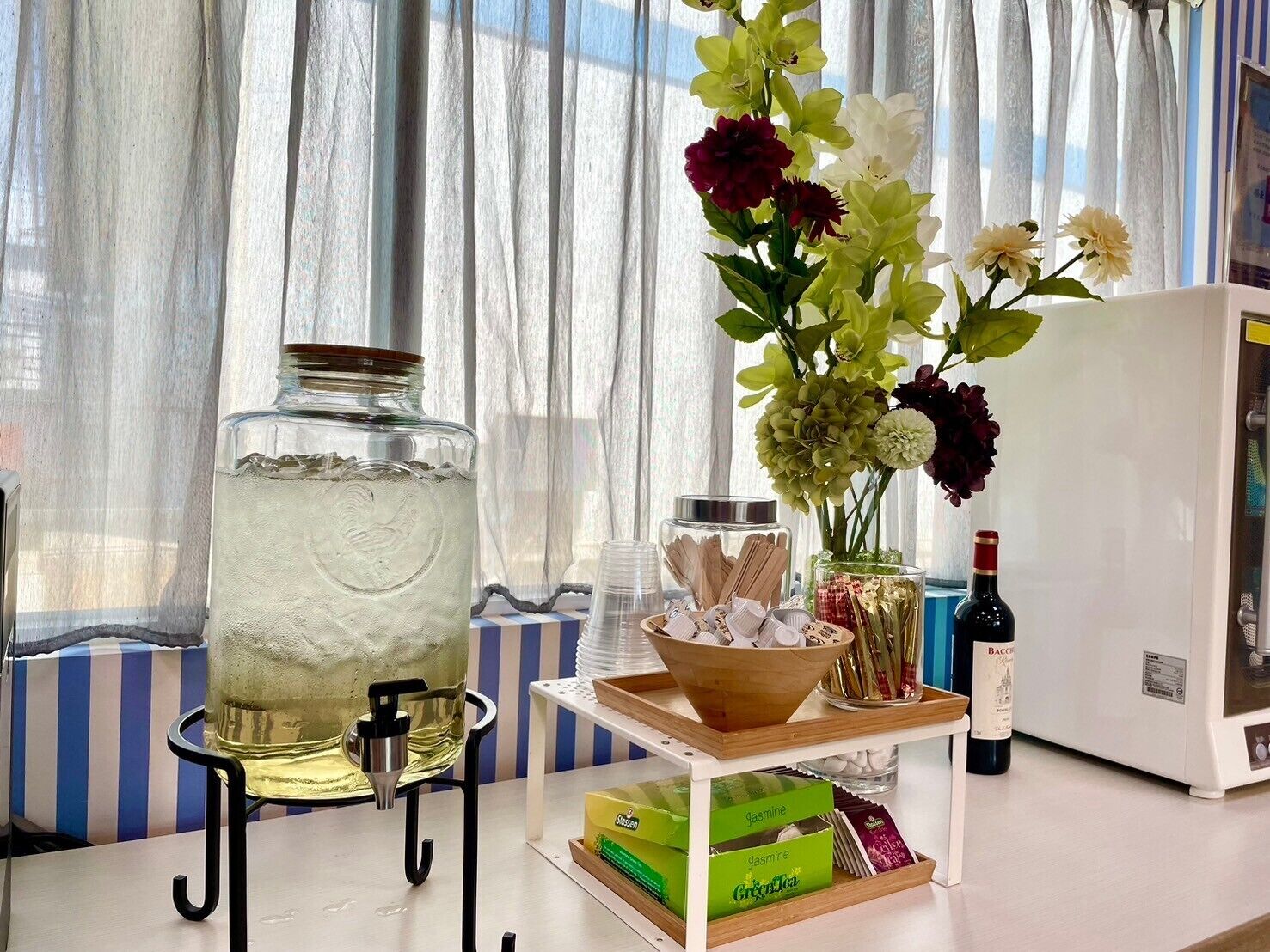 A kitchen counter with a large glass jar, a bottle of wine, a vase with flowers, and a bowl of cookies.