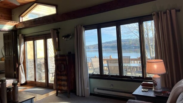 Une pièce chaleureuse avec une grande fenêtre donnant sur un lac et des montagnes. On y trouve un ventilateur de plafond, une commode en bois et une lampe sur une table d’appoint.