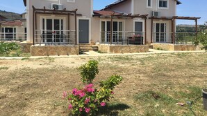 Ein zweistöckiges Haus mit Steinfundament, einer überdachten Terrasse und einem kleinen Garten mit rosa Blumen.