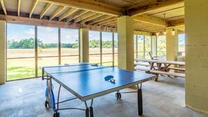 A ping pong table in a room with large windows, wooden beams, and picnic tables.