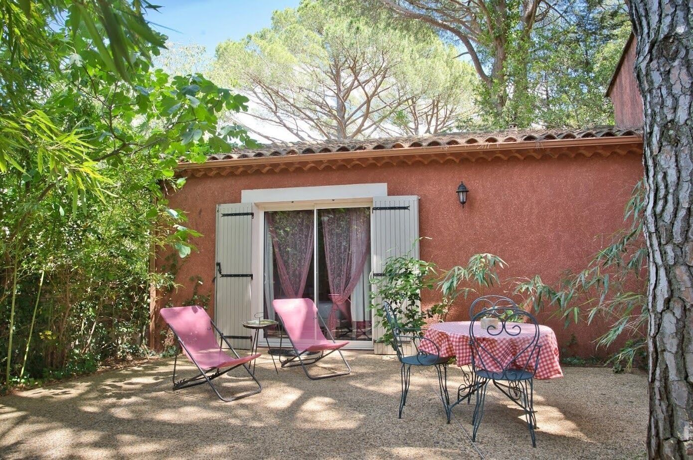 Un patio con dos sillas rosadas y una mesa con mantel a cuadros, rodeado de follaje y un edificio con tejado de tejas.