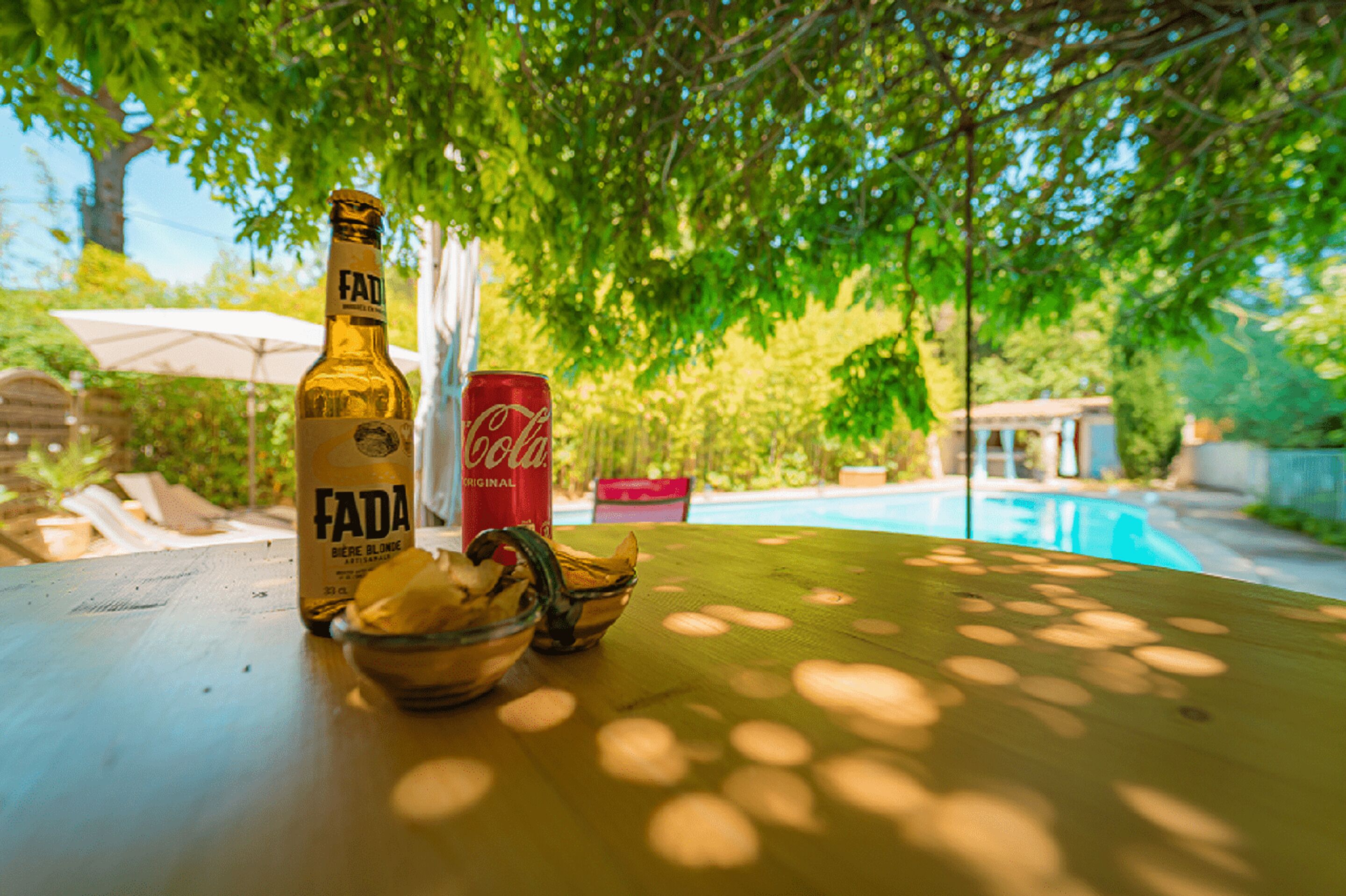 A bottle of Fada beer, a can of Coca-Cola, and a bowl of fruit on a table by a poolside.