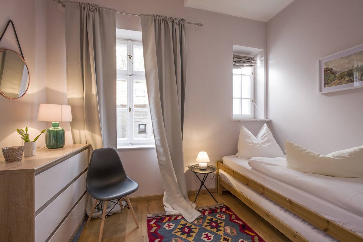 Un dormitorio con cama, escritorio con lámpara, silla, mesita con lámpara y ventana con cortinas.